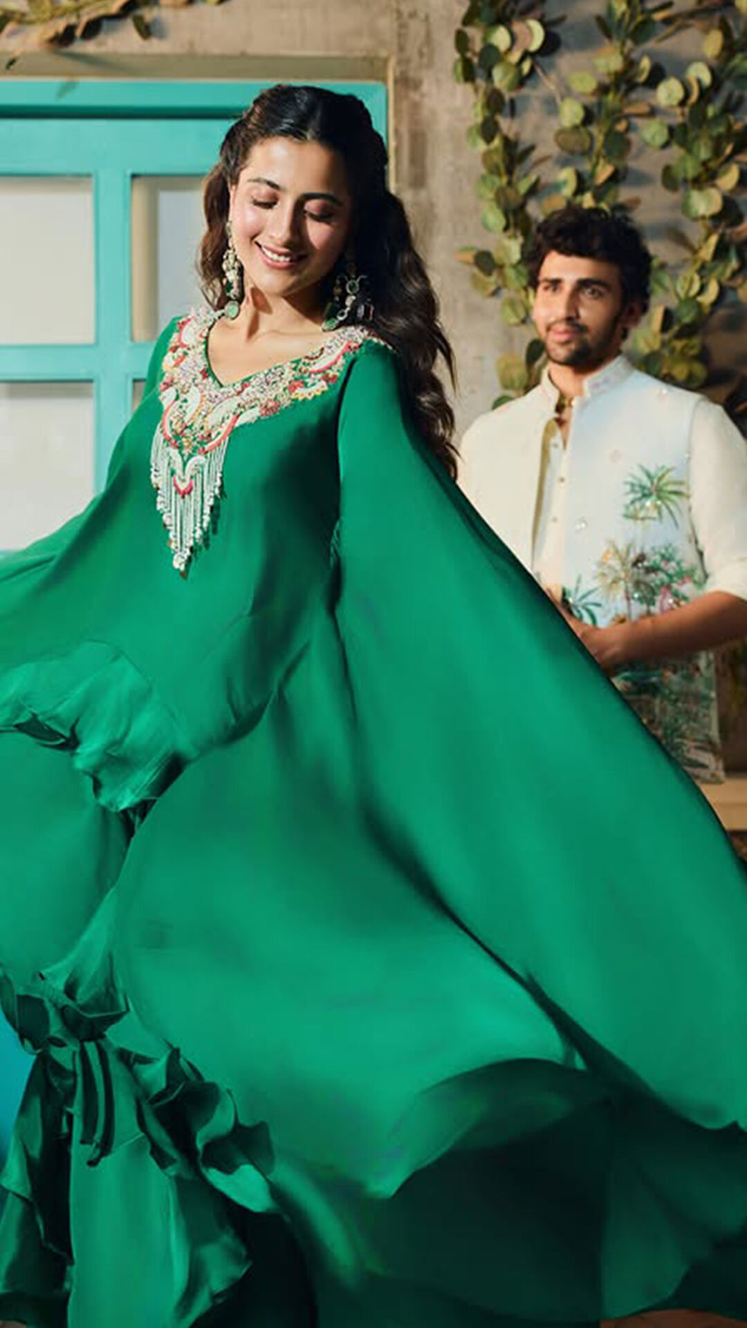 Woman in an emerald green flowing anarkali with mirror work, standing next to a man in an embroidered jacket, highlighting festive wear.